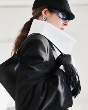 Profile of woman in black hat and sunglasses wearing an oversized jacket with a high white collar and carrying a fringe handbag.