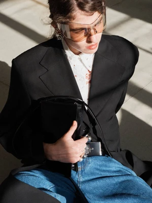 Model wearing a black blazer, white shirt, blue jeans, and visor sunglasses holding a black handbag.