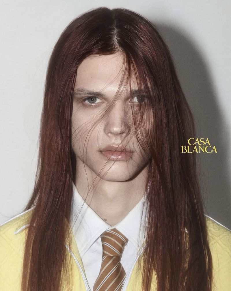 Portrait of a male model with long auburn hair wearing a white shirt and striped tie against a neutral background.