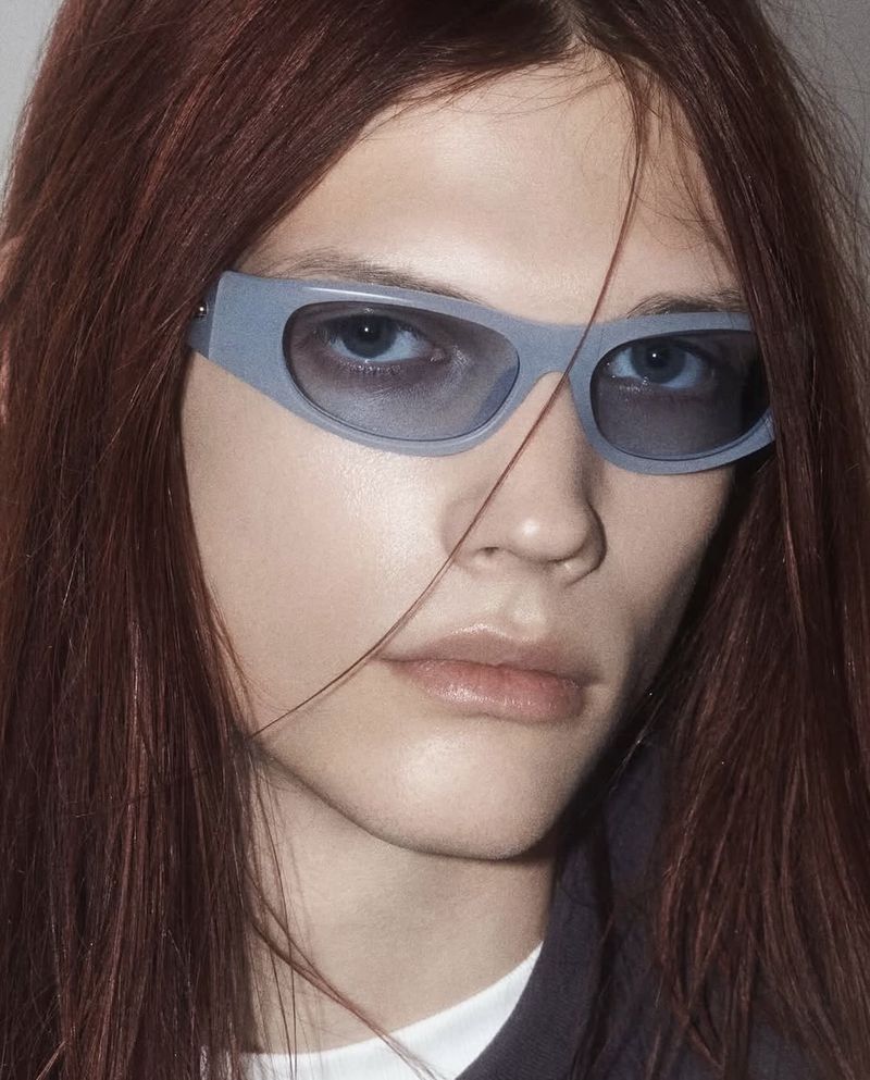 Close-up of a person with auburn hair wearing grey sunglasses. The shot focuses on their face and the eyewear.