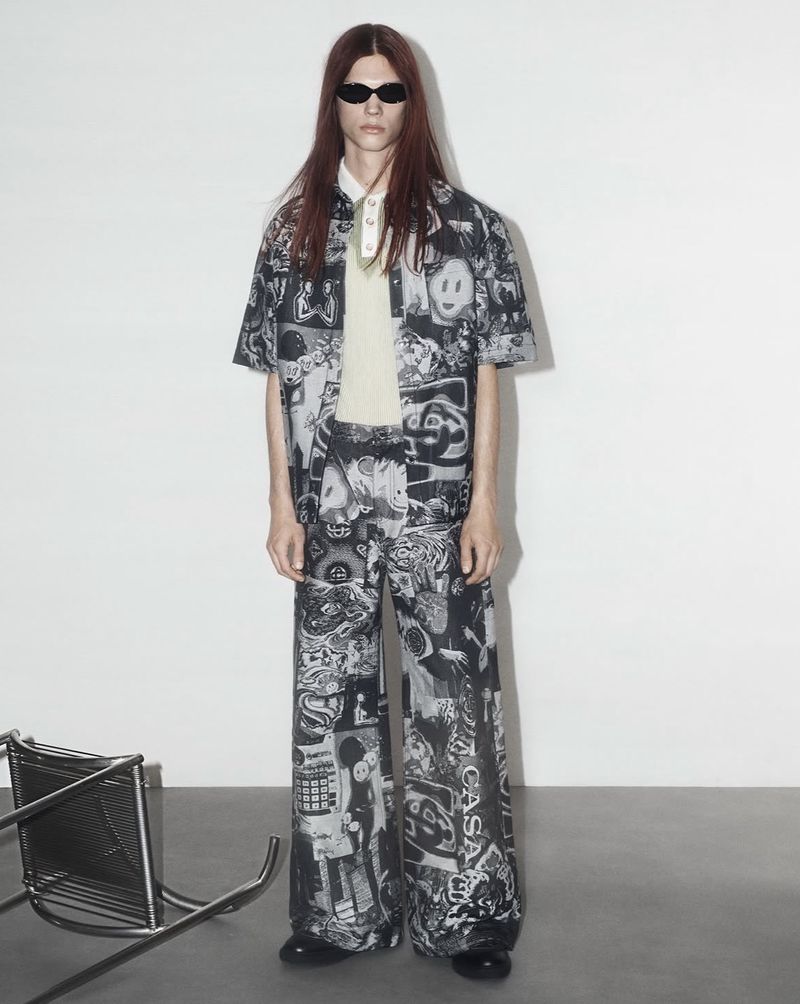 Model wearing dark sunglasses and a graphic print shirt and trousers set standing against a plain white studio backdrop.