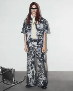 Model wearing dark sunglasses and a graphic print shirt and trousers set standing against a plain white studio backdrop.