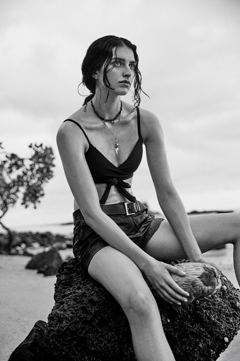 Model Saffron Vadher wearing a black crop top and belted shorts seated on a coastal rock holding a coconut in black and white.