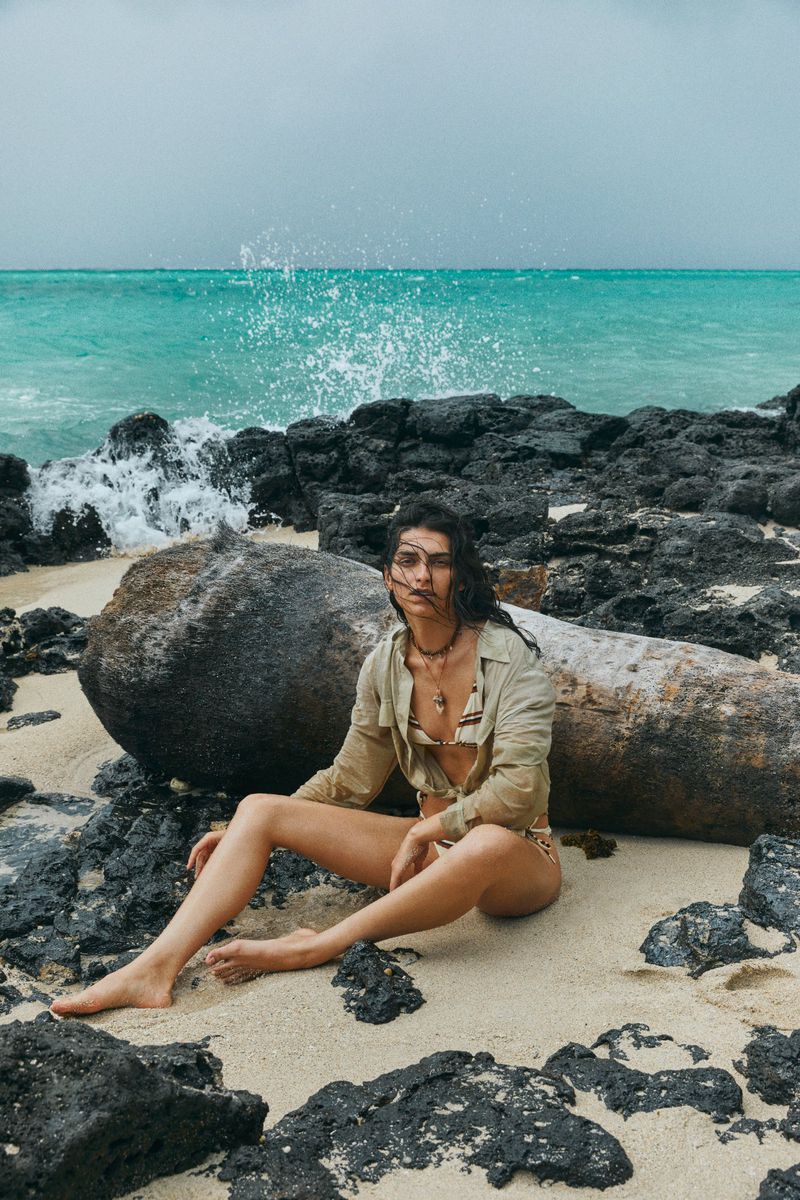 A woman in swimwear and an unbuttoned shirt sits on a sandy beach among rocks and driftwood, with the ocean and sky in the background.