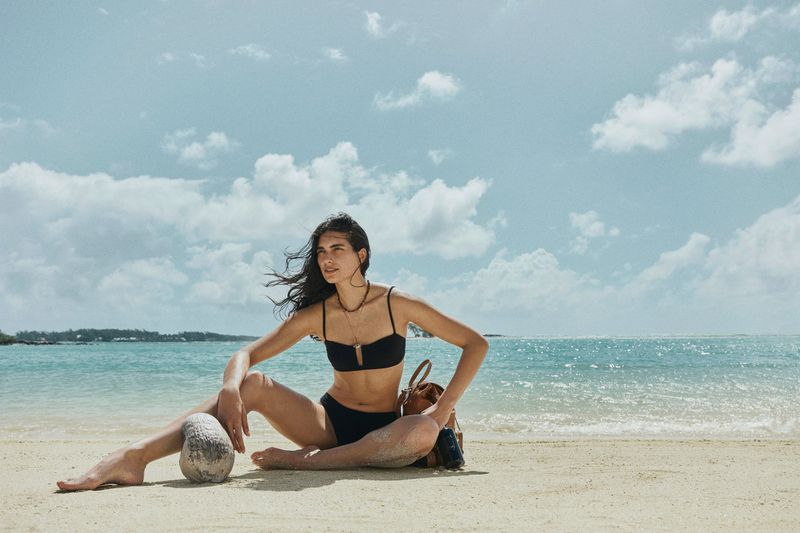 A tanned woman in a black bikini sits on a sandy beach next to a coconut. The ocean and a cloudy sky are visible in the background.