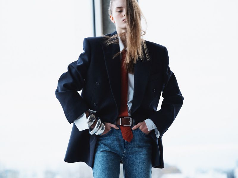 Model in oversized black blazer wearing a white shirt, red tie, and blue jeans against a bright window.