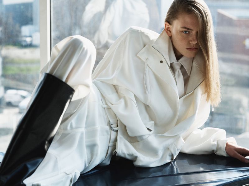 Model wearing a white jacket and matching pants with black boots reclining indoors near a large window for The Frankie Shop campaign.
