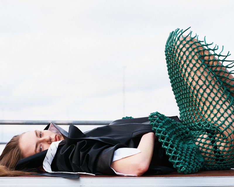Model lying on a surface wearing a dark jacket and a green netted skirt against an overcast sky