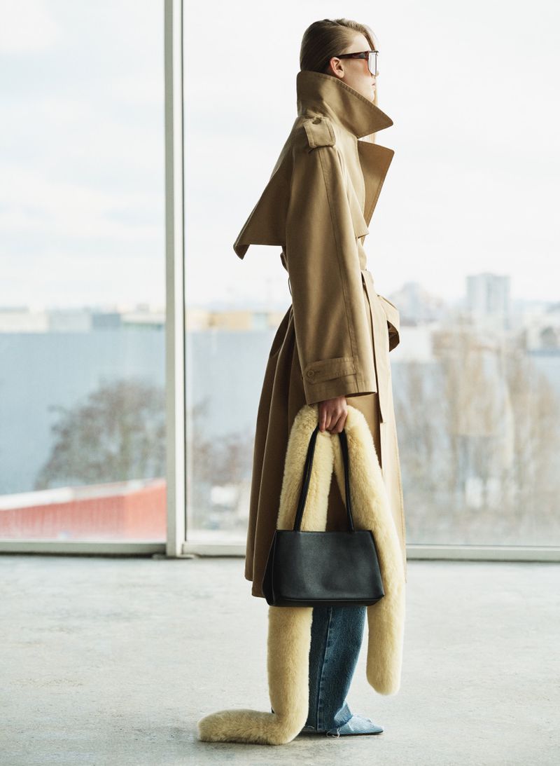 Model in camel trench coat and sunglasses holding a black handbag inside near a large window