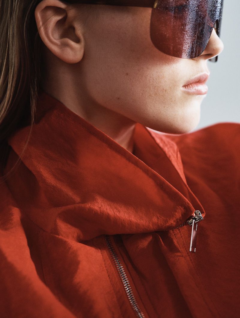 Close-up portrait of a model wearing dark oversized sunglasses and a red zip-up jacket against a light grey background.