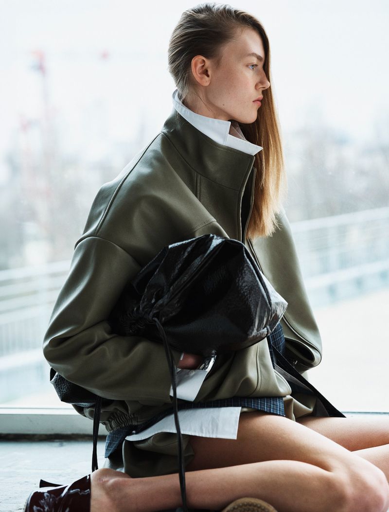 Model in olive green jacket wearing a white collared shirt holding a dark handbag for The Frankie Shop campaign