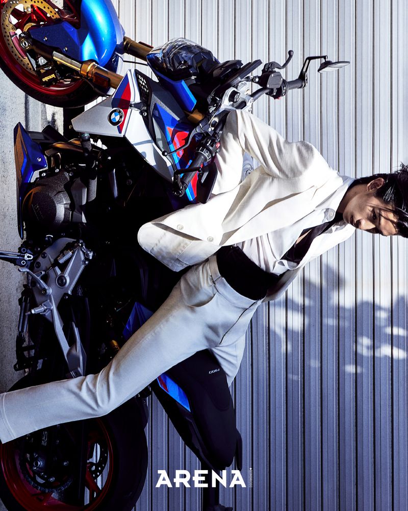 Model wearing a white suit posing beside a blue and white motorcycle against a vertical metal shutter background.