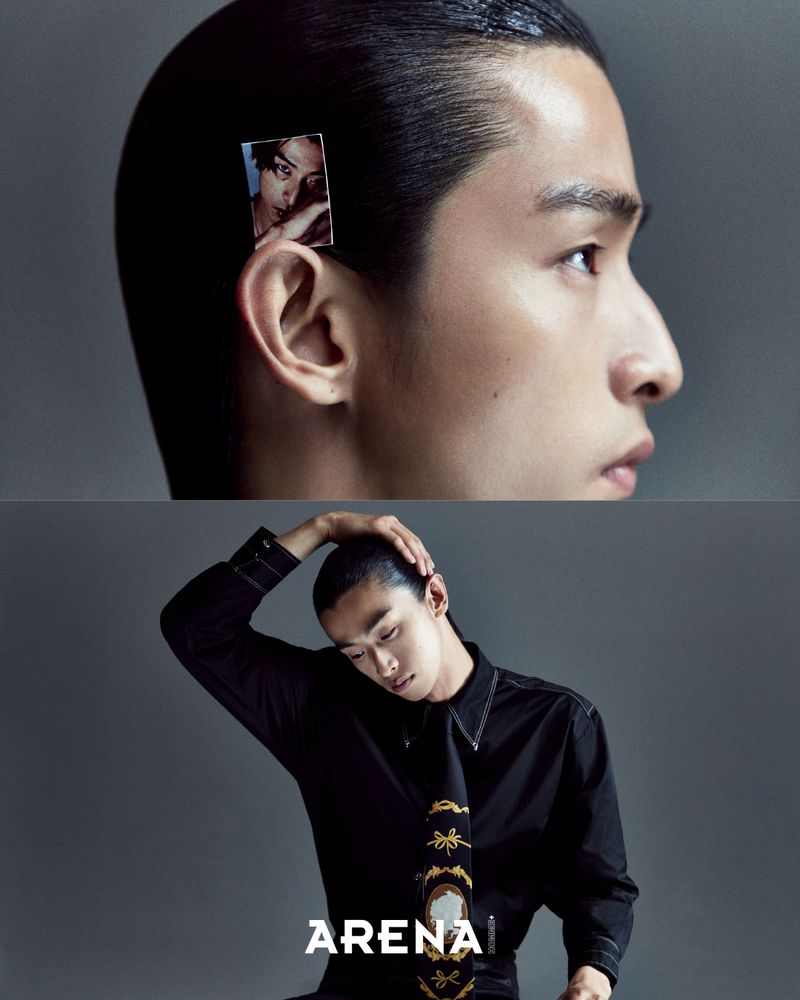Close-up portrait of a male model with slicked-back dark hair and a small printed photograph pinned near his ear against a grey background.