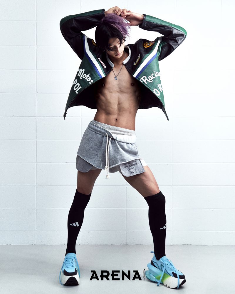 Model with purple-streaked hair wearing a green leather racing jacket, grey shorts, knee-high socks, and blue sneakers against a tile background.