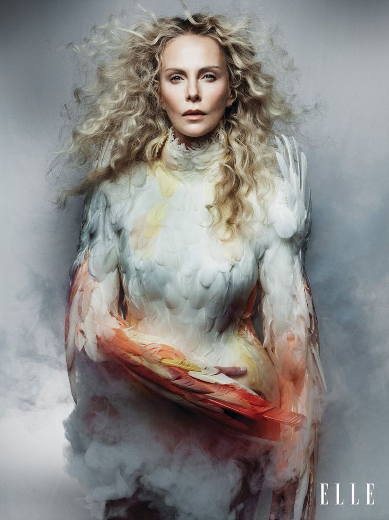 Charlize Theron wearing a feathered gown with voluminous blonde curly hair against a smoky grey studio backdrop.