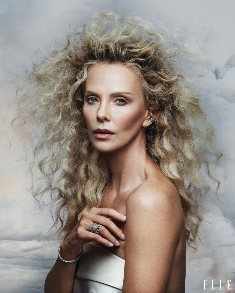 Charlize Theron with voluminous blonde hair against a cloudy sky backdrop for Elle U.S. portrait.