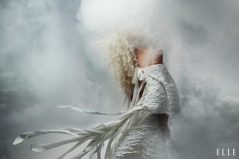 Charlize Theron from behind with voluminous curly blonde hair in a textured white dress surrounded by clouds of smoke.