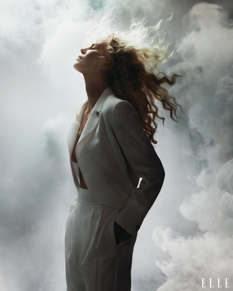 Elegant woman in white suit with flowing blonde hair poses in a surreal, smoky studio setting for an Elle magazine fashion editorial.