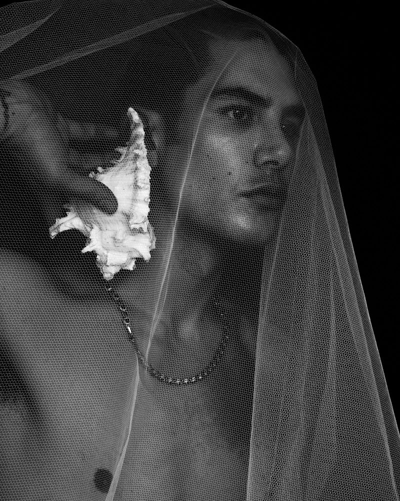 Close-up portrait of a man draped in sheer white tulle holding a white seashell to his ear against a solid black background.
