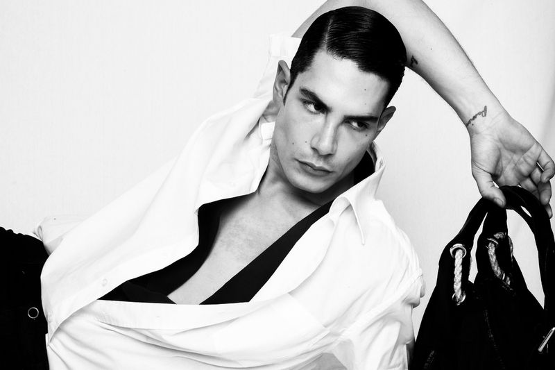 Monochrome fashion portrait of a man with slicked-back hair in an open white shirt holding a black bag against a white backdrop.