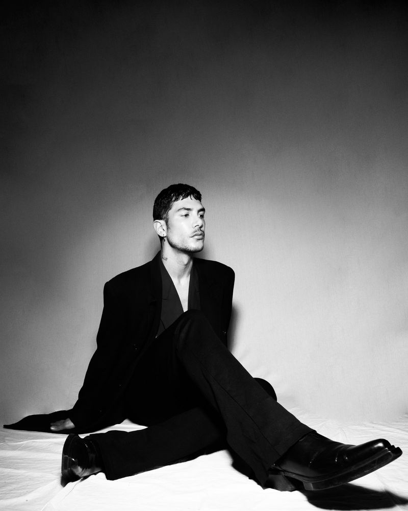Monochrome studio portrait of a man in a black suit sitting on a light backdrop for Mirror Mirror Magazine.