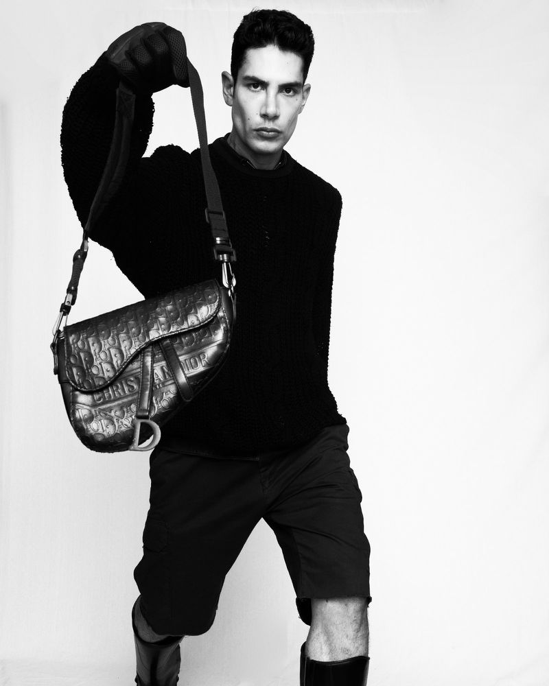 Male model in an all-black outfit posing with a black leather saddle bag against a clean white studio backdrop.