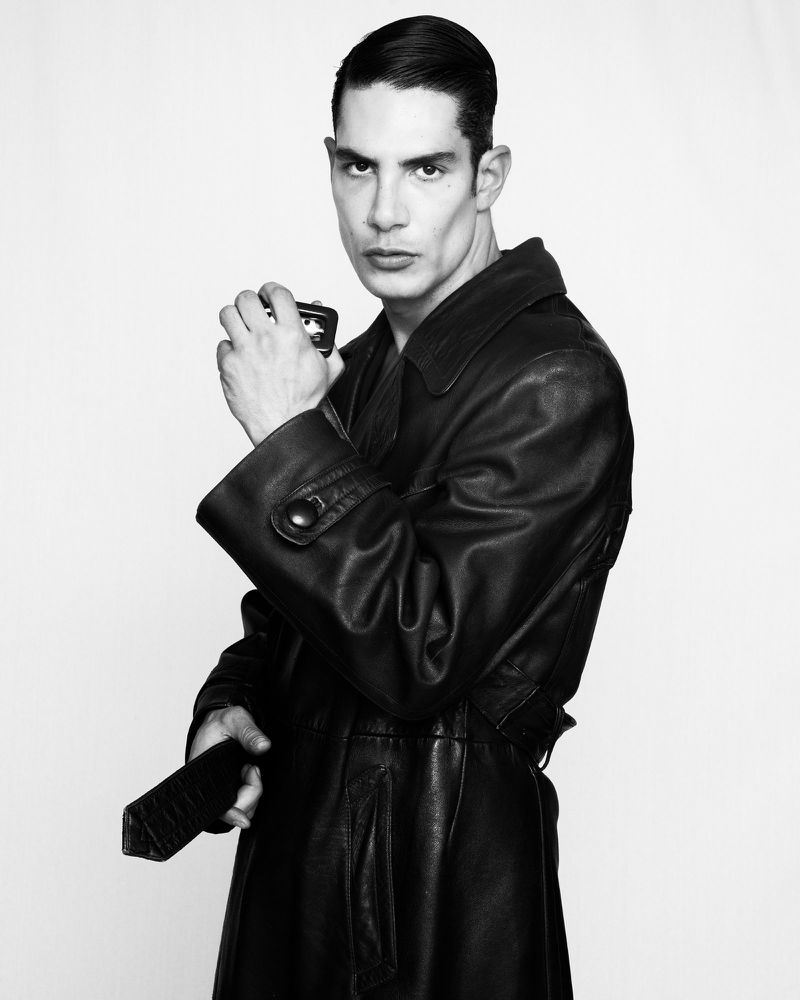Monochrome portrait of a man with slicked hair wearing a dark leather trench coat against a bright white background.