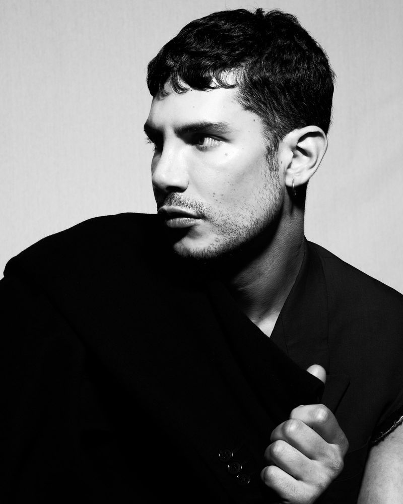 Black and white portrait of a man looking to the side wearing a dark structured jacket against a plain backdrop.