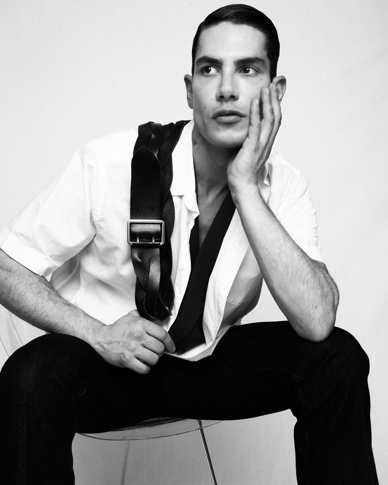 Man in a white shirt and black trousers wearing a braided leather harness seated against a white backdrop.