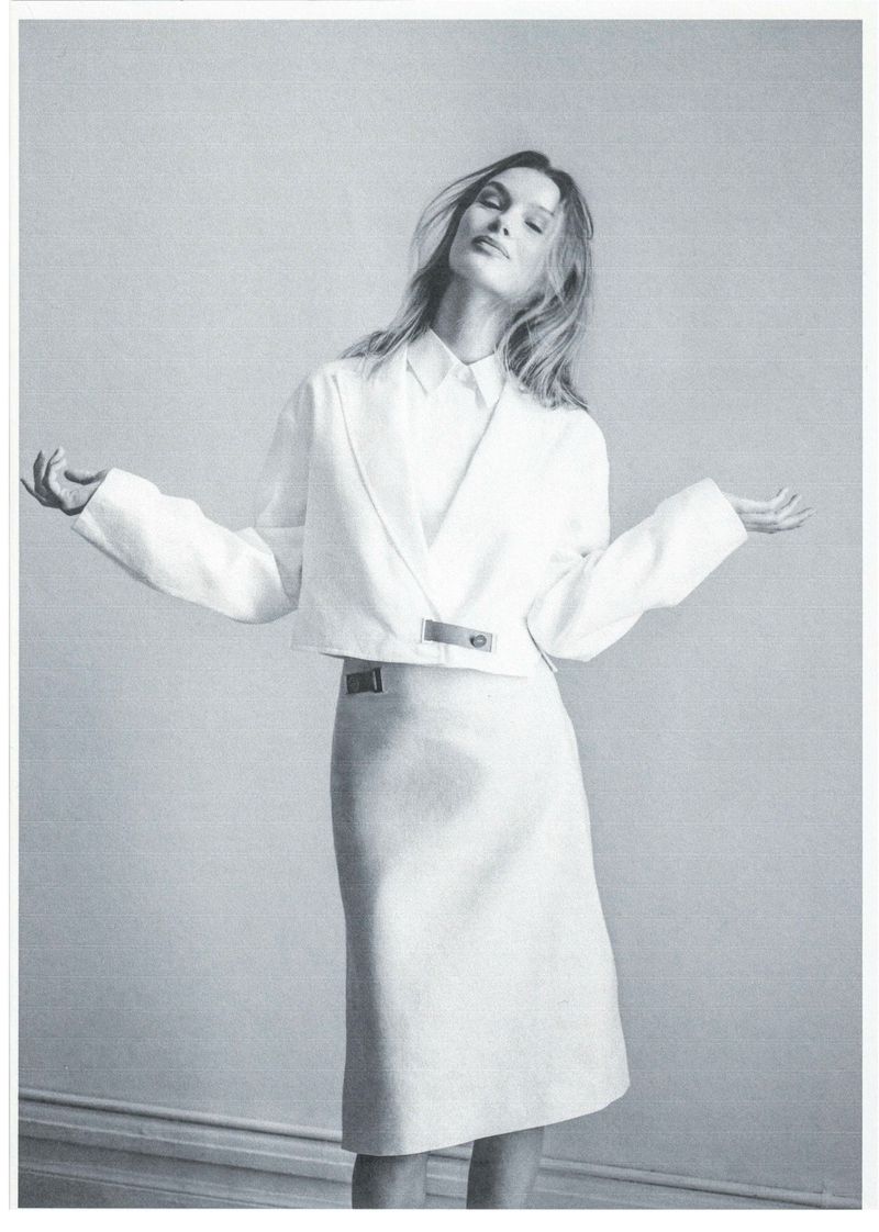 Model Kris Grikaite posing in a white cropped blazer and matching midi skirt against a light studio wall.