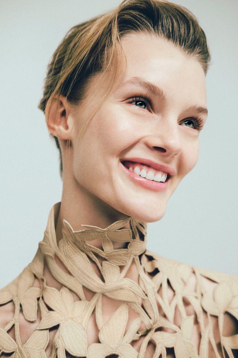 Kris Grikaite smiling in a beige laser-cut floral high-neck garment against a pale studio backdrop.