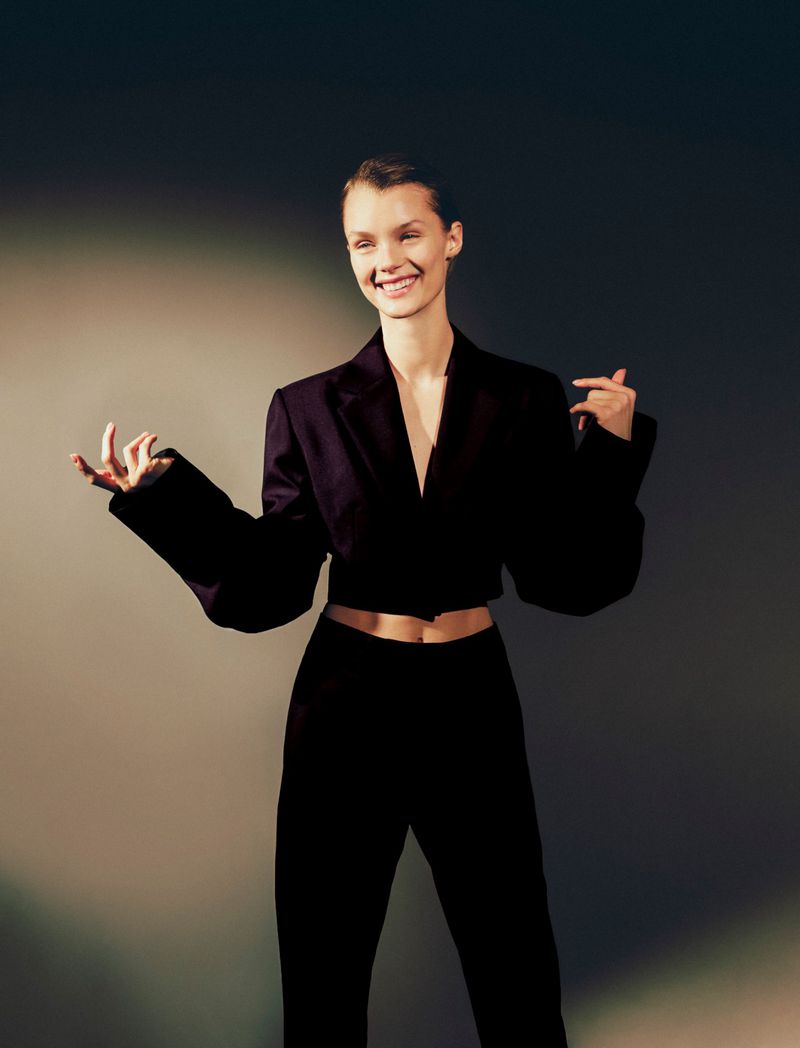 Model Kris Grikaite smiling in a black cropped blazer and matching trousers against a studio gradient backdrop.