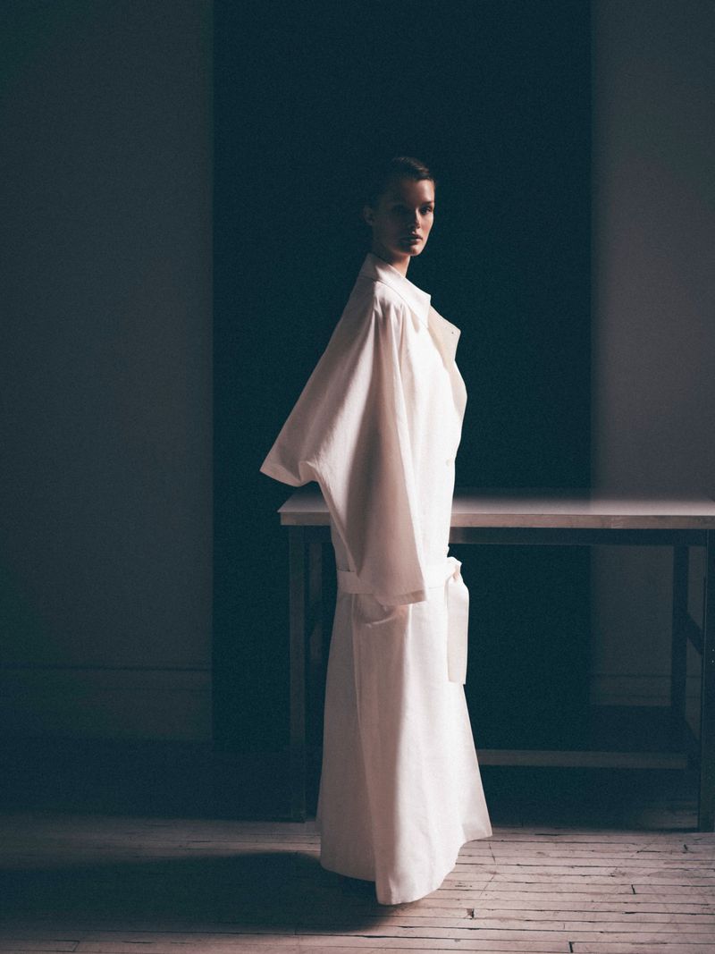 Model Kris Grikaite posing in an oversized white belted robe coat against a dark backdrop in a studio.