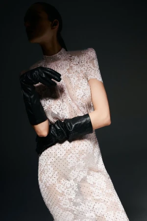 Model wearing a pale sheer floral lace high-neck dress and black leather opera gloves against a dark background.