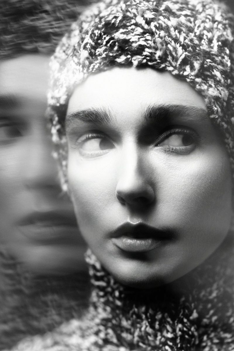 Close-up black and white portrait of a person wearing a textured knit cap and sweater with a double exposure blur effect.