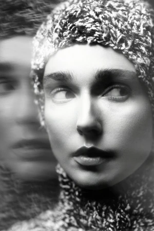 Close-up black and white portrait of a person wearing a textured knit cap and sweater with a double exposure blur effect.