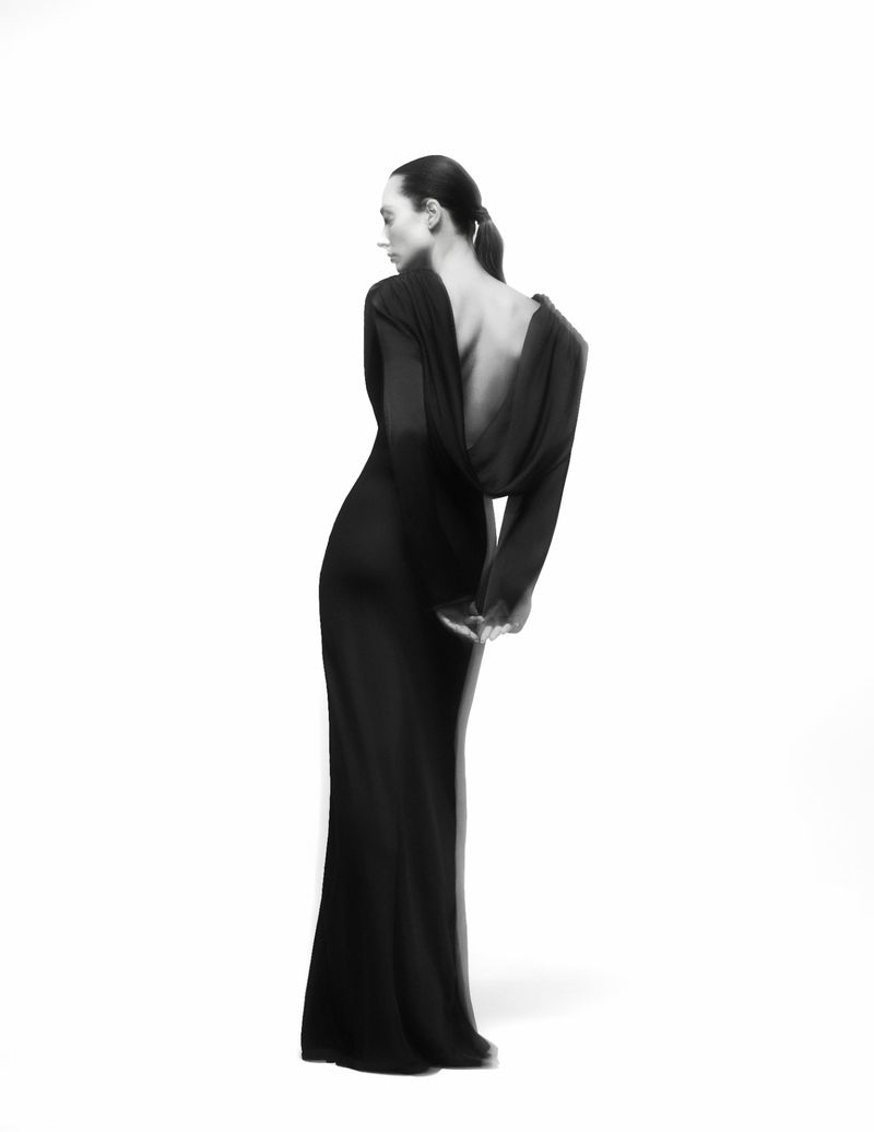 Woman with a sleek low ponytail posing in a long black backless gown against a clean white studio backdrop.