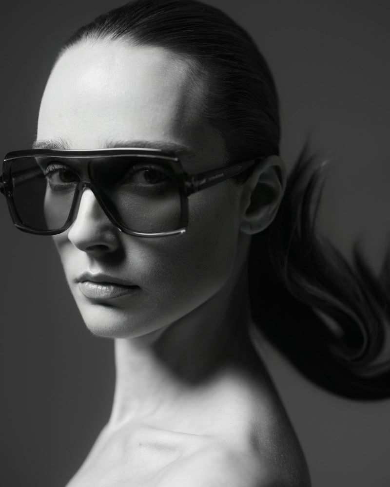 Close-up portrait of a woman wearing oversized sunglasses with her hair styled in a ponytail against a plain background