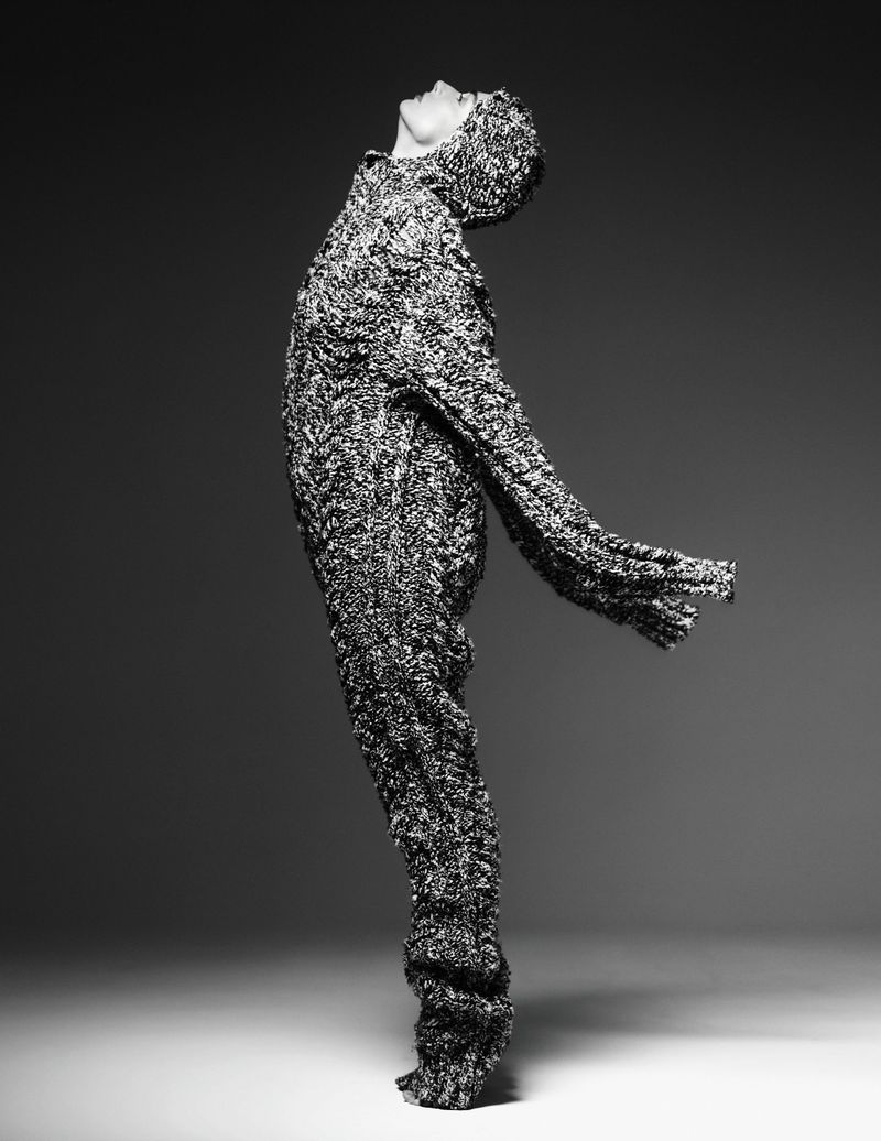 A full-length studio shot in black and white of a person in a textured long-sleeved outfit with a hood, leaning their head back.