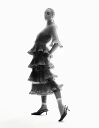Model wearing a layered tiered dress and slingback heels against a bright white background in a fashion editorial shot.