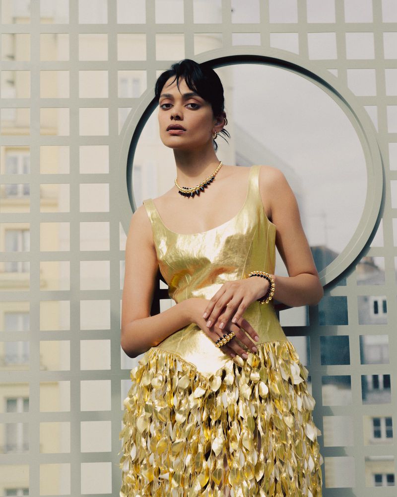 Model Zinnia Rose Kumar wearing a metallic gold dress and statement jewelry outdoors against a geometric grid screen.