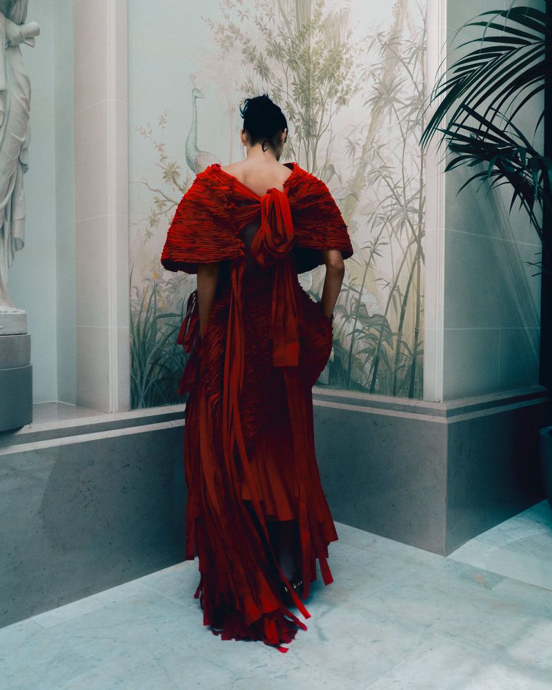 Model Zinnia Kumar seen from behind wearing a sculptural textured red gown and cape against an interior botanical painted wall.