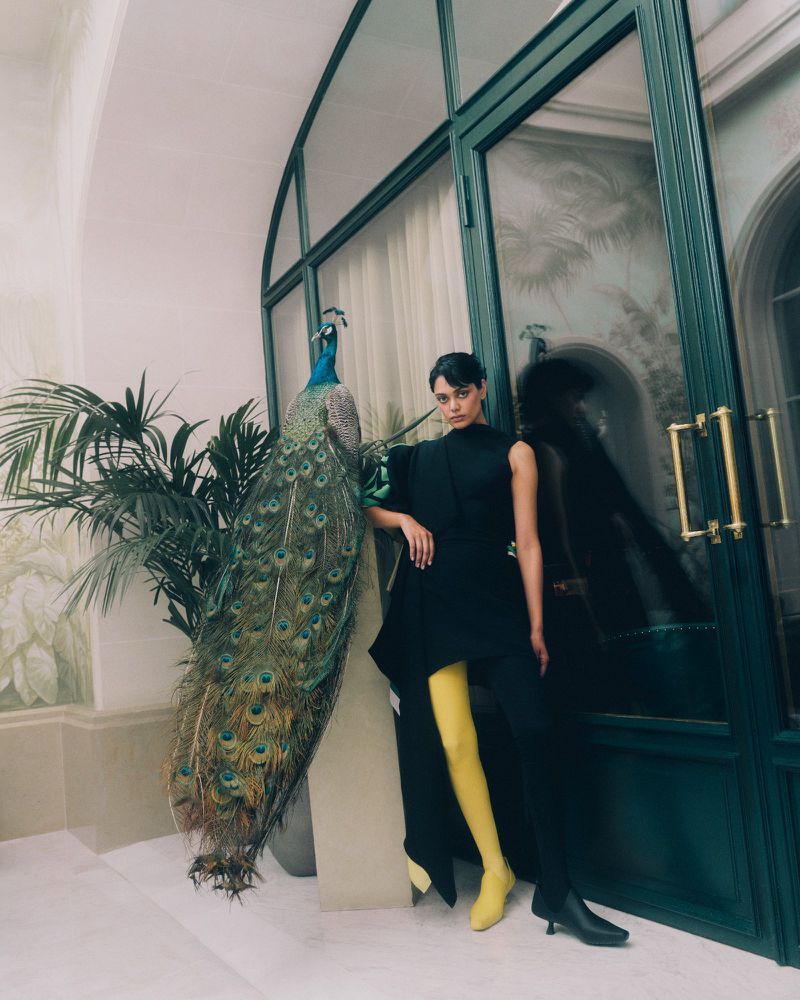 Model in black top and yellow tights posing next to a taxidermy peacock near a dark green glass door