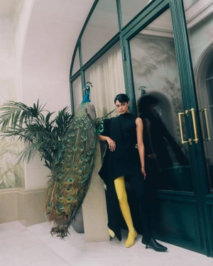 Model in black top and yellow tights posing next to a taxidermy peacock near a dark green glass door