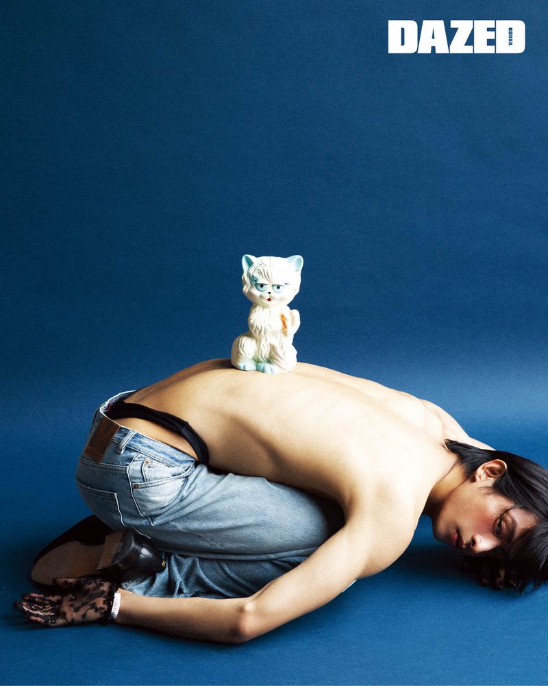 Model Yugo Takano crouched shirtless in denim jeans with a ceramic cat figurine on his back against a blue studio backdrop.