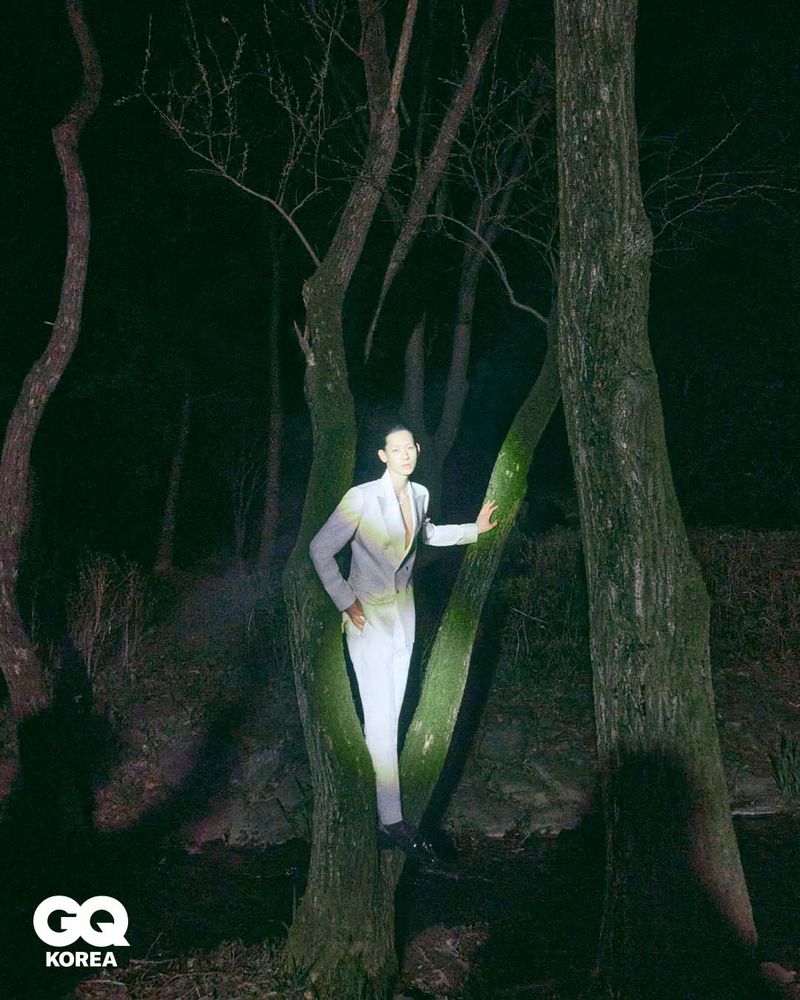 Model wearing a light suit standing between dark trees at night with a spotlight illuminating them for GQ Korea.
