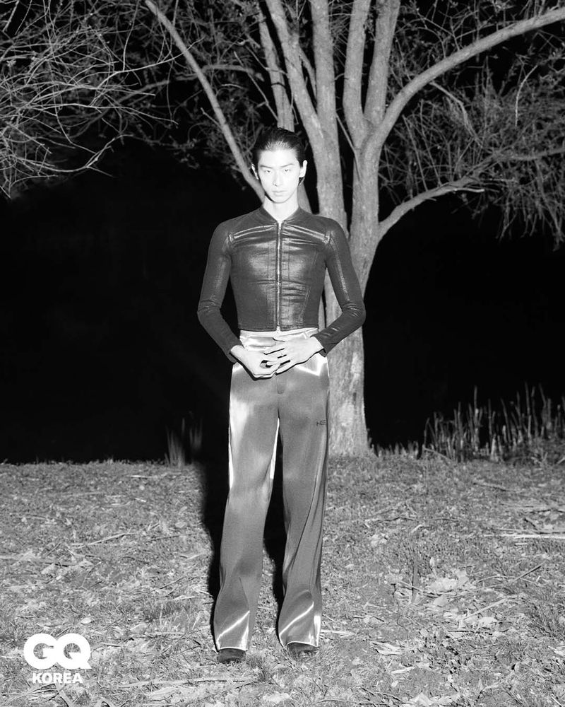 Model Yugo Takano in a dark fitted jacket and glossy trousers standing outdoors at night in front of a tree for GQ Korea.