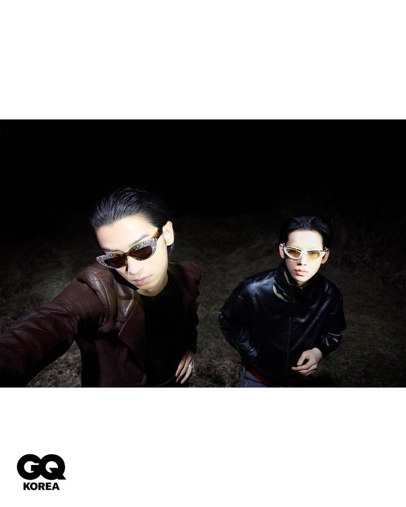 Two men wearing sunglasses and dark jackets posing outdoors at night for GQ Korea under directional flash lighting