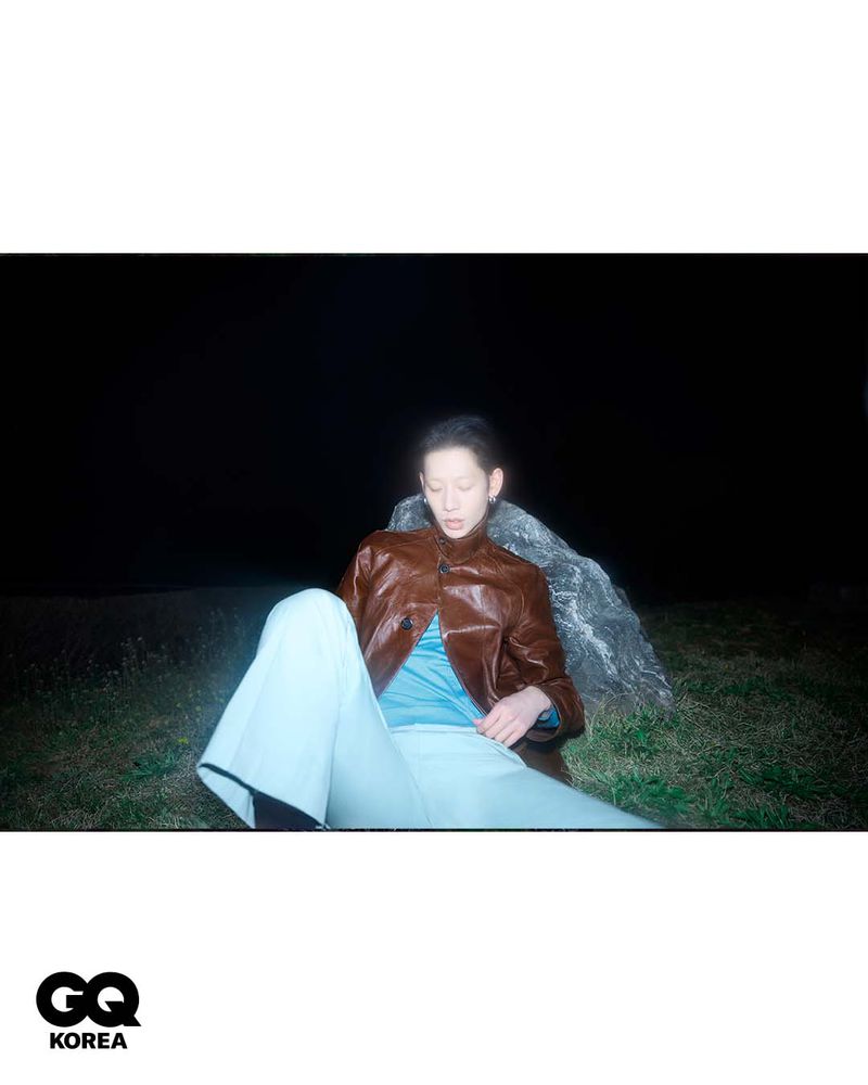 Model Yugo Takano seated on grass leaning against a rock at night wearing a brown jacket and light blue trousers.
