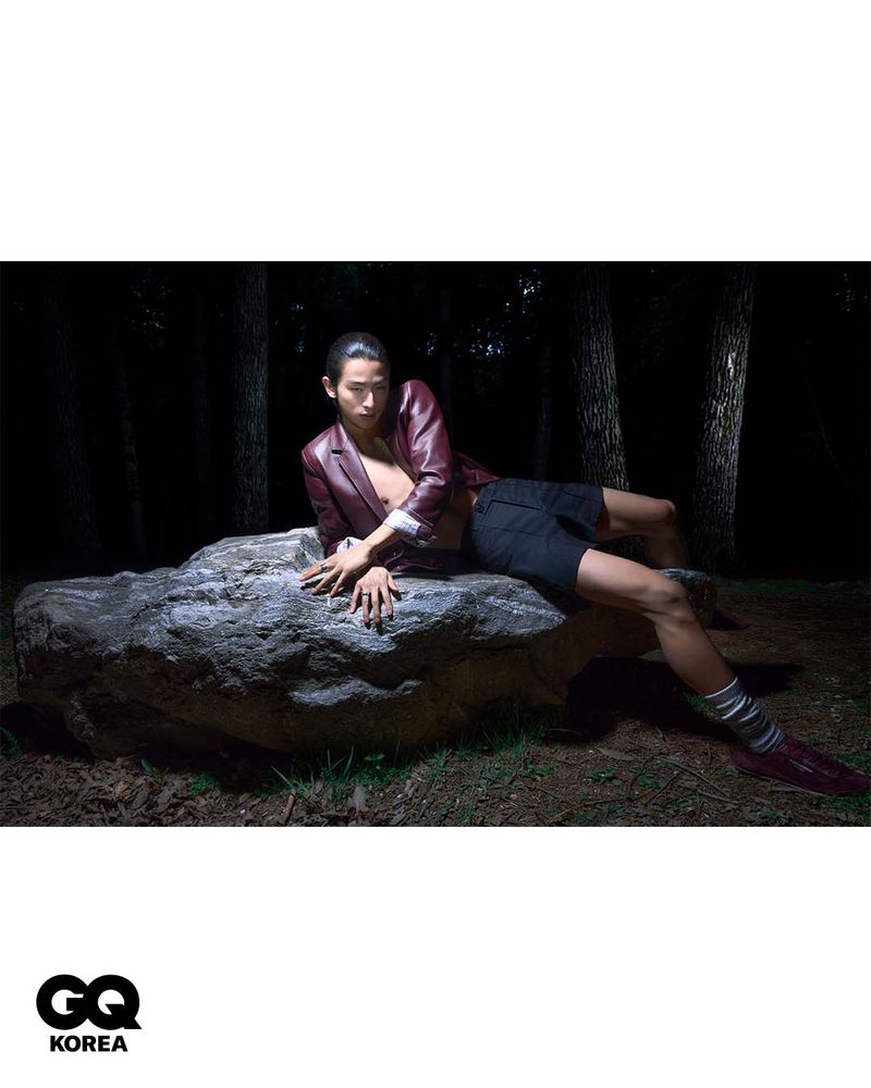 Model wearing a burgundy leather jacket and black shorts reclining on a large stone in a dark wooded setting.